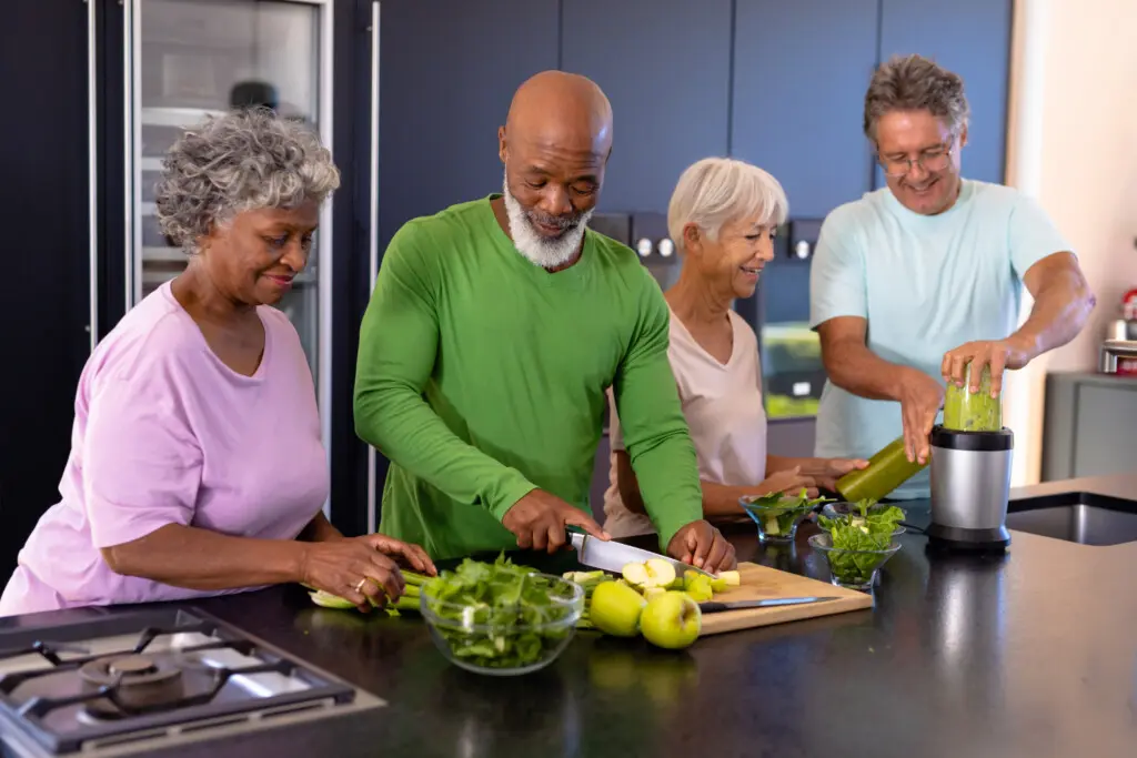 Multiracial seniors friends chopping fruits, vegetables and blending smoothie on kitchen counter. Retirement home, unaltered, togetherness, support, assisted living and healthy food concept.